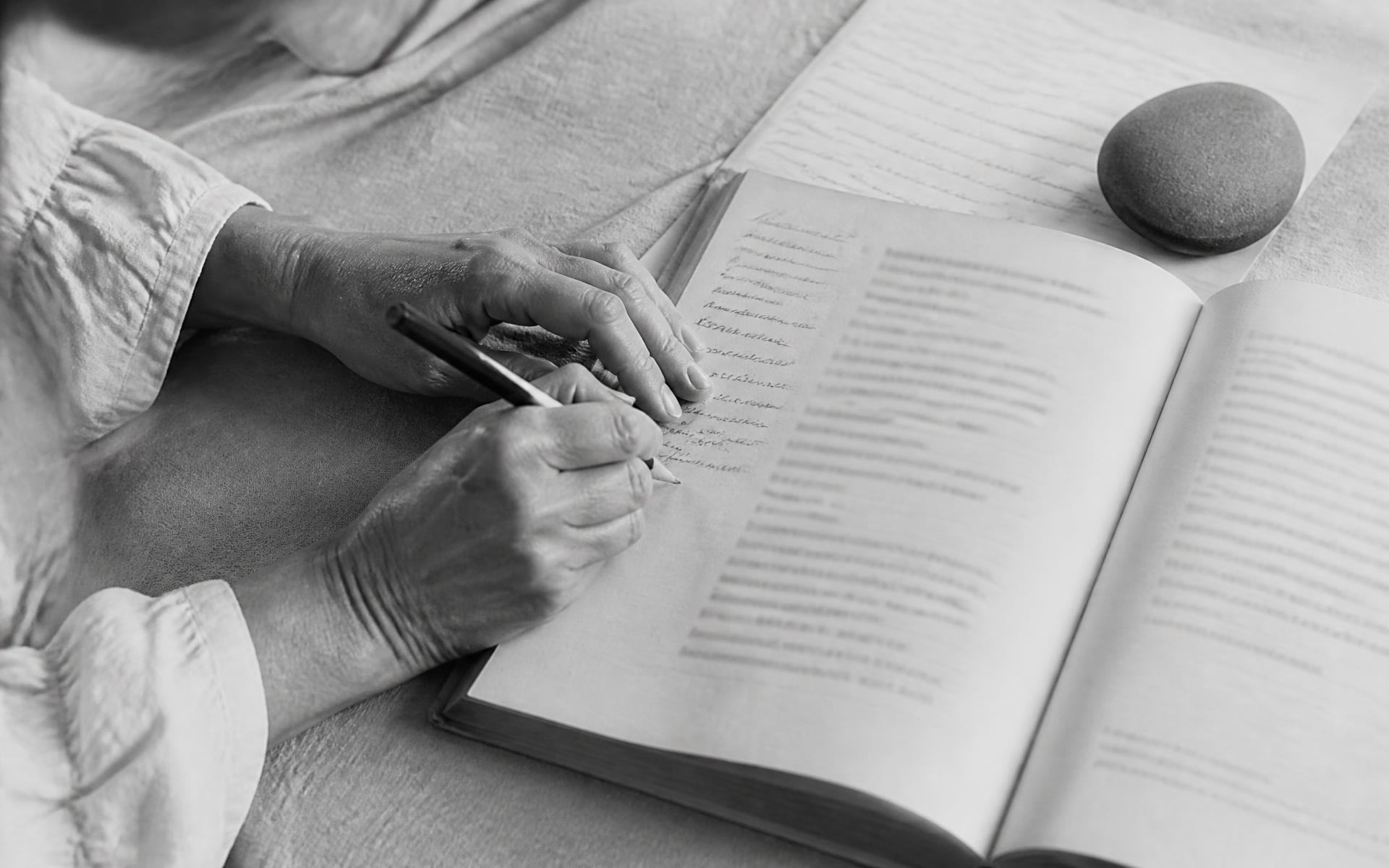 Hands annotating an open workbook with a pencil beside a stone paperweight